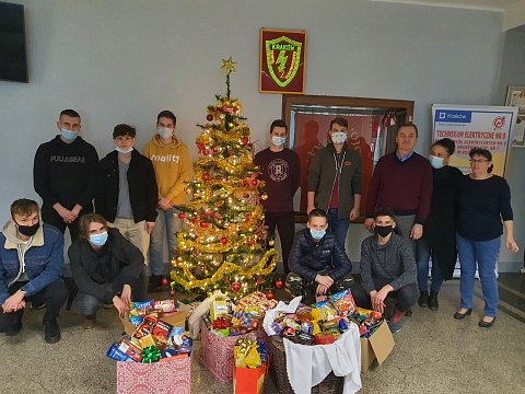 Uczniowie i nauczyciele w maseczkach stoją obok choinki. Przed nimi kosze z darami