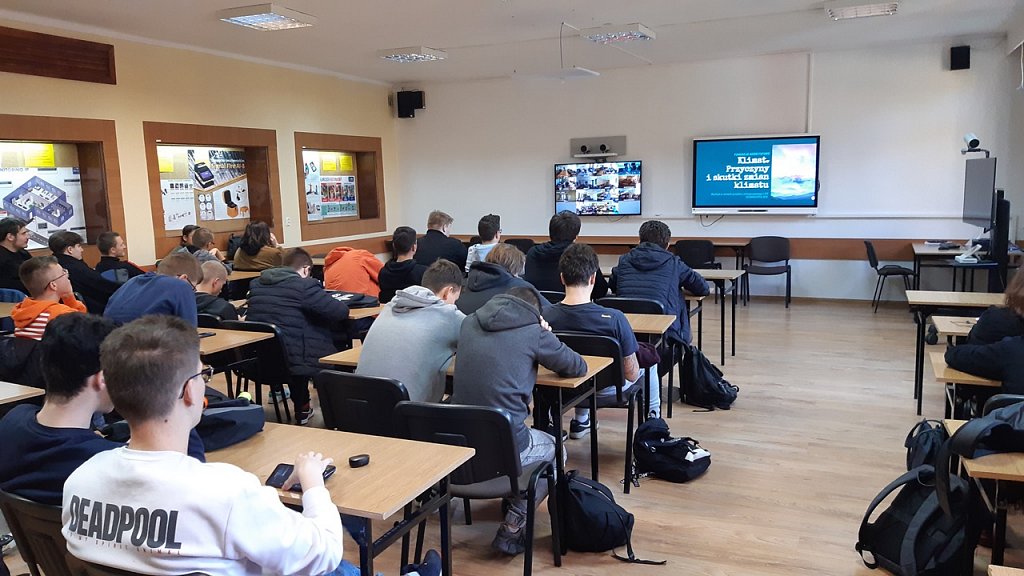 Zdjęcie sali konferencyjnej. Na ścianie wisi duży telewizor. Uczniowie w ławkach oglądają transmisję wykładu.