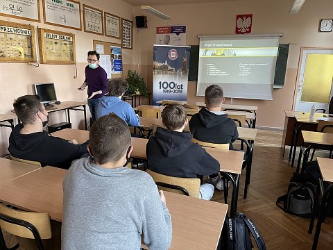Uczniowie w sali lekcyjnej z uwaga słuchają wykładu prowadzonego przez wykładowcę w maseczce.