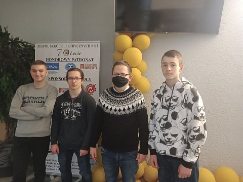 Trzech uczniów z nauczycielem stoi w holu szkoły. W tle banery i baloniki - dekoracja z okazji 70 lecia szkoły. 