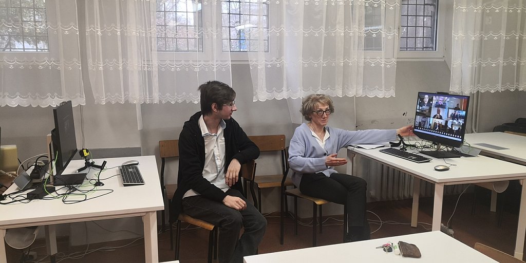 Dwa stoliki z ustawionymi przy nich monitorami. Przy jednym siedzi uczeń, przy drugim nauczyciel. 