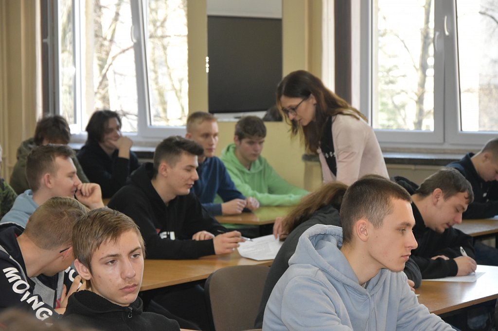 Sala lekcyjna. Uczniowie siedzą  w ławkach. Stojący nauczyciel rozdaje testy.