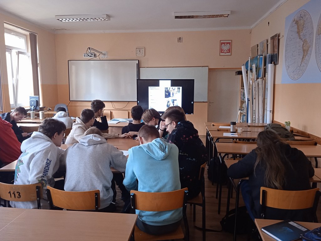 Sala lekcyjna. uczniowie pracują w grupach. Na telewizorze stojącym obok ekranu wyświetlona prezentacja.