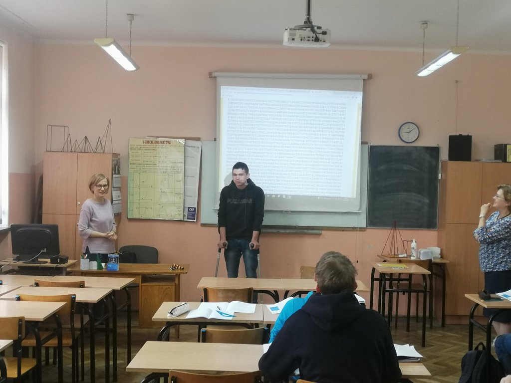 Sala lekcyjna. Pod ekranem z wyświetloną prezentacja stoi uczeń ( opiera się na kulach). w ławkach siedzą uczniowie. Stoi dwóch nauczycieli.