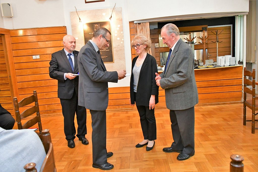 Saka konferencyjna. W tle bufet. Panowie w garniturach wręczają odznaczenie.