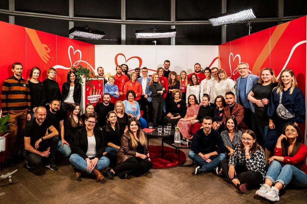 Studio telewizyjne. Grupa uczestników. W tle białe i czerwone banery. 