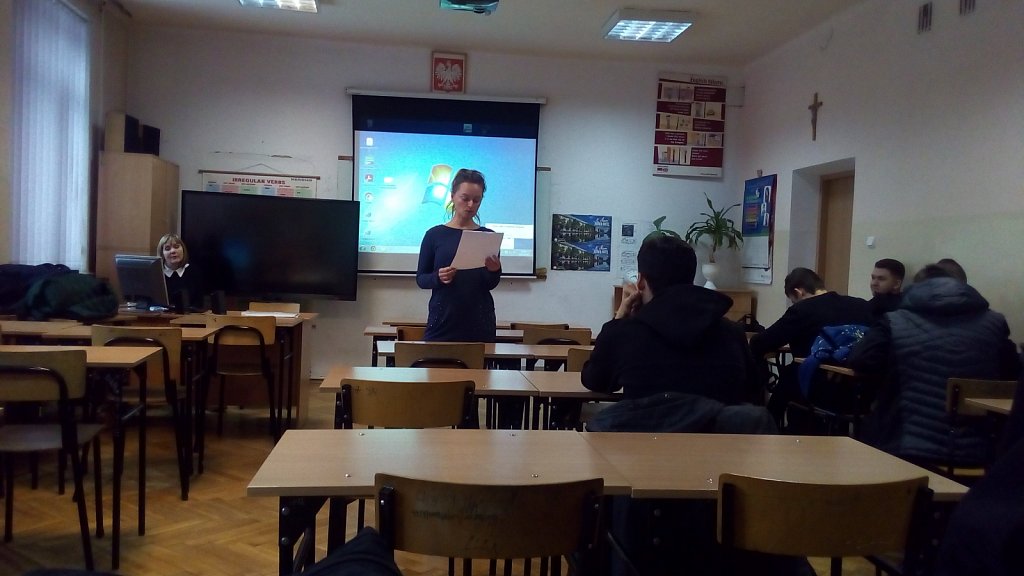 Sala lekcyjna. W tle ekran z prezentacją. Grupa uczniów siedzi w ławkach. Nauczyciel stoi do nich przodem. W ręce trzyma kartkę. 