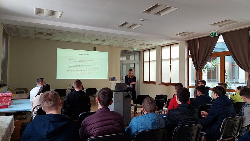 Sala konferencyjna z dużymi oknami. Na ekranie wyświetlona prezentacja. Uczniowie słuchają wykładu. 